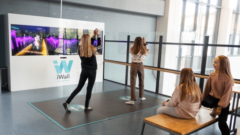 Two girls interact with a dance game on a large screen, while two others watch. The setup is labeled iWall.